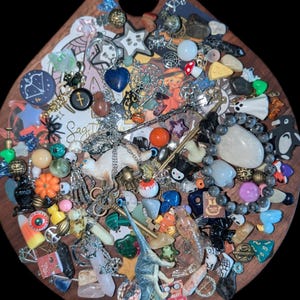 May include: A teardrop-shaped wooden board covered in a collection of small charms, beads, crystals, and miniature figurines, including a dinosaur, a cat, and a penguin. The text "Sagittarius" and "Large Witchy Mystery Scoop" are visible.