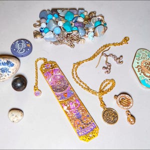 May include: A collection of Scorpio-themed items including a purple and gold metal bookmark, a light green enamel pin with a scorpion graphic and the text Scorpio, a gold chain necklace with a circular pendant, earrings, loose stones, and beads.