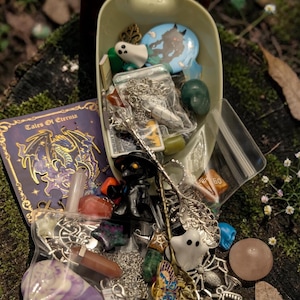 May include: A selection of fantasy-themed charms, pendants, and small figurines, including a ghost, a black cat, and a butterfly, spilling from a light green scoop. A purple book with gold lettering and the text "Tales Of Eternia" is visible.