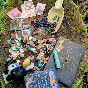 Pode incluir: Uma coleção de itens místicos, incluindo um baralho de cartas de oráculo de sereias, um diário com um desenho de sol, cristais, amuletos e um brinquedo de pelúcia, dispostos em um toco de árvore coberto de musgo. Uma concha contém velas.
