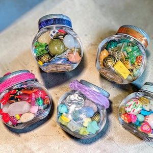 May include: Five small, clear glass jars filled with assorted colorful beads, charms, and stones. The jars have metal lids, some wrapped with pink or purple yarn, and others featuring decorative patterns like stars or anchors on the rim.