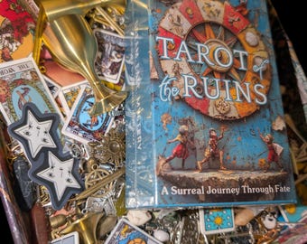 Puede incluir: Una caja que contiene una baraja Tarot of the Ruins en un empaque azul, dos cálices de metal dorado, varias cartas de tarot, pegatinas en forma de estrella con caras y diversos pequeños dijes de metal y cristales esparcidos.