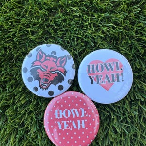 May include: Three round buttons with graphic designs. One button features a red wolf head on a white background with black polka dots. Another has "HOWL YEAH!" with a red heart. The third button has "HOWL YEAH!" on a red polka dot background.