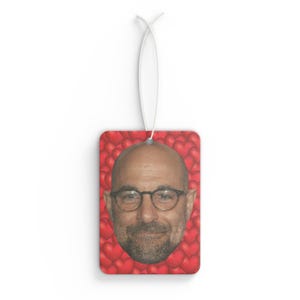 May include: Rectangular air freshener featuring a man's face with glasses, set against a background of red hearts. The air freshener is suspended by a white string.