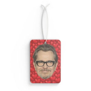Puede incluir: Ambientador rectangular con el retrato de un hombre con gafas, sobre un fondo de corazones rojos. El ambientador tiene una cuerda blanca para colgar.