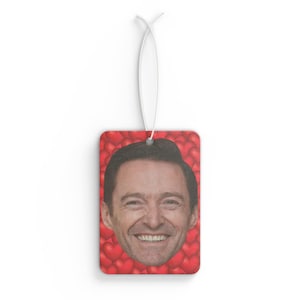 Puede incluir: Ambientador rectangular con una cara sonriente sobre un fondo de corazones rojos. Tiene una cuerda blanca para colgar. El diseño está impreso en una forma rectangular.