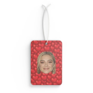 Puede incluir: Ambientador rectangular con un fondo de corazones rojos y la cara de una mujer en el centro. El ambientador está colgado por un cordón blanco. El fondo es un patrón de corazones rojos.