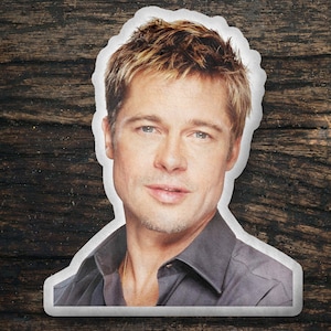 May include: A close-up portrait of a man with short, light brown hair. He is wearing a dark gray shirt and has a gold chain necklace around his neck.