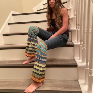 May include: A person wearing blue jeans and a black tank top with colourful crocheted leg warmers. The leg warmers have a striped pattern with brown, green, blue, and orange colours.