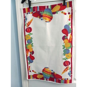 May include: A white rectangular kitchen towel featuring a colorful border pattern of stylized pottery, round fruits, and feathers in shades of red, yellow, blue, and green. The design frames a central white space, finished with a striped red edge.