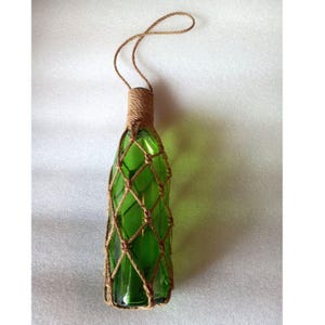 May include: A green glass bottle encased in a brown jute rope netting that covers the body and wraps around the neck. The rope extends upward from the bottle neck to form a loop for hanging, creating a decorative vessel with a textured finish.