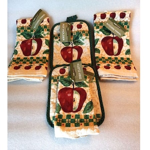 May include: A kitchen set featuring a red apple design on a cream background. The set includes two kitchen towels, a pot holder, and a dish cloth. Each item has a green border and a tag that reads "Kitchen Towel", "Pot Holder", or "Dish Cloth".