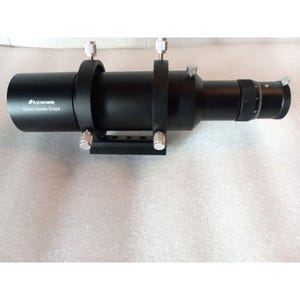 May include: A black Astromania 60mm guide scope featuring a cylindrical body, two mounting rings with silver adjustment screws, and a focus tube with a graduated scale. The device is designed for astronomical observation and telescope alignment.