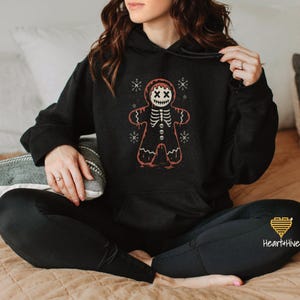Peut inclure: Sweat à capuche noir avec un motif de squelette de bonhomme en pain d'épices. Le bonhomme en pain d'épices a des yeux "XX" et une cage thoracique de squelette. Le motif est rouge et blanc, avec des flocons de neige autour.