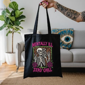 May include: Black tote bag featuring a skeleton with the words "MENTALLY ILL" and "ZERO CHILL" in pink and green. The skeleton is making a peace sign and holding a phone. The design includes roses and a leopard print background.