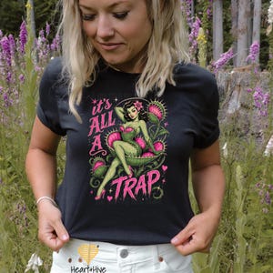 May include: Black t-shirt featuring a pin-up style illustration of a woman in a Venus flytrap. The design includes the text "It's All A Trap" in pink and green. The woman is depicted with green skin and pink accents.