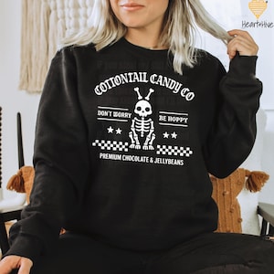 Può includere: Felpa nera con grafica bianca raffigurante un coniglio scheletro e la scritta "COTTONTAIL CANDY CO". Il design include le frasi "DON'T WORRY" e "BE HOPPY", insieme a "PREMIUM CHOCOLATE & JELLYBEANS".