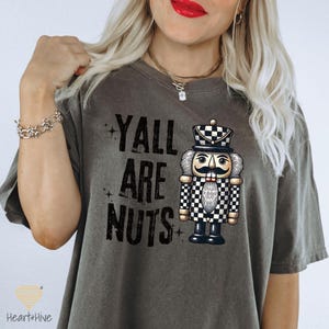 May include: Dark grey t-shirt with the text "YALL ARE NUTS" and a graphic of a nutcracker soldier. The nutcracker has a black and white checkered uniform and a large mustache. The shirt is a comfortable, casual style.