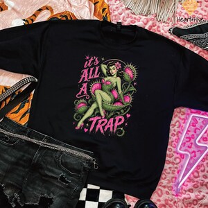 May include: Black sweatshirt featuring a pin-up style illustration with the text "it's ALL A TRAP". The graphic depicts a woman in a Venus flytrap setting, with pink and green accents. The sweatshirt is paired with black denim shorts and accessories.