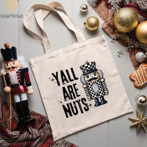 May include: A cream-colored tote bag with the text "Y'ALL ARE NUTS" and a cartoon nutcracker illustration. The nutcracker is in a black and white checkered uniform. The bag has two cream-colored handles.