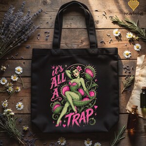 May include: Black tote bag featuring a pin-up style illustration of a woman with green skin and a pink corset, seated in a Venus flytrap. The text "it's ALL A TRAP" is displayed in pink. The bag has black handles.