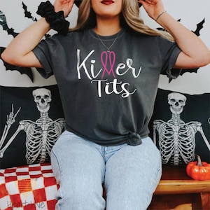 May include: Dark gray t-shirt with the words "Killer Tits" in white script and a pink ribbon design. The shirt is knotted at the waist. The background includes Halloween decorations and skeleton pillows.