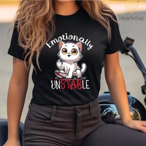以下が含まれることがあります： 血まみれのナイフを持った白い漫画の猫が描かれた黒いTシャツ。Tシャツには「感情的に不安定」という文字が印刷されています。