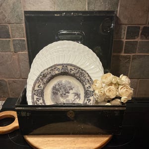 May include: A black metal box with a stack of white and blue patterned plates. A small bouquet of cream-colored flowers sits to the right. The plates have a detailed design. The box is on a wooden surface.