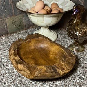 May include: A rustic, wooden bowl with a natural, irregular edge. The bowl has a warm, brown color and a smooth interior. A decorative bowl with a checkered pattern and a gold rim holds several brown eggs. A gold-colored glass rooster figurine is also present.