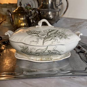 May include: A white porcelain serving dish with a lid, featuring a floral design in shades of gray and green. The dish sits on a matching oval-shaped plate, with a silver tray in the foreground. Other silver serving pieces are in the background.
