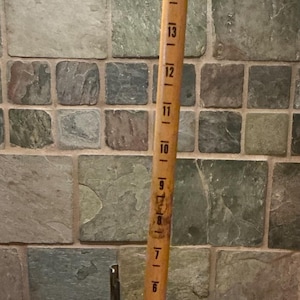 May include: A vintage Dritz skirt marker, a measuring tool for sewing, stands on a metal base. The wooden ruler is marked with inch increments from 6 to 18. The tool is set on a wooden surface.