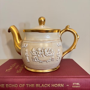 May include: A cream-colored teapot with a gold-painted spout, handle, lid knob, and base rim. The body features a raised white relief scene of figures seated around a table. The teapot rests on a red book titled The Echo of the Black Horn by Ethel Knight.