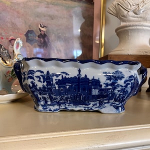May include: A rectangular ceramic planter featuring a blue and white transferware pattern depicting a classical architectural scene with buildings, trees, and figures. The vessel has a scalloped top edge, two side handles, and a footed base.