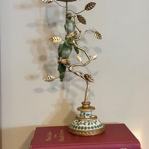 May include: A decorative candle holder featuring a gold-toned metal tree branch with two green parrot figurines perched on it. The base and top candle cup are white porcelain with green leaf patterns and gold trim. It stands on a red book titled The Echo of the Black Horn.