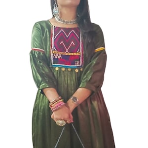 May include: A green long-sleeved dress with embroidered details and gold buttons. The dress has a gathered skirt and is paired with black pants and gold shoes. The model is holding a beaded bag with a fringe.