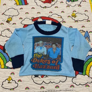 Peut inclure: Un t-shirt à manches longues bleu clair avec un col et des poignets bleu marine. Le t-shirt présente une image vintage des personnages des Dukes of Hazzard, Bo et Luke Duke, avec le titre de la série en lettres orange et jaunes. Le t-shirt porte un avis de copyright datant de 1971.