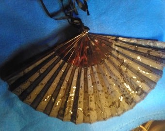 May include: A vintage hand fan with a black and gold design. The fan features a pleated, semi-transparent material with black lace accents and gold glitter. A black ribbon is attached at the top. The fan is open, displaying its full shape.