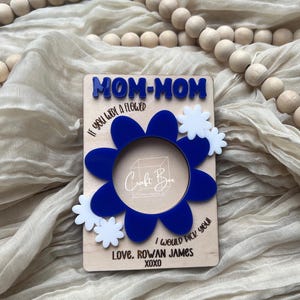 May include: A wooden card with the text "MOM-MOM" in blue letters. The card has a blue flower with a white center and white flowers around it. The text "IF YOU WERE A FLOWER I WOULD PICK YOU! LOVE, ROWAN JAMES XOXO" is written on the card.