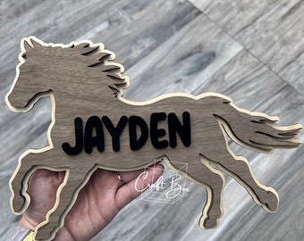 May include: Wooden horse silhouette with the name "JAYDEN" cut out in black lettering. The horse is brown and has a mane and tail. The background is a wood grain pattern.