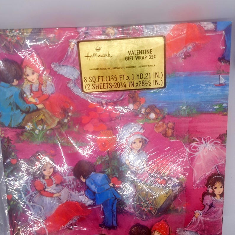 May include: A package of Hallmark Valentine gift wrap featuring a pink background with illustrations of children in vintage clothing, hearts, and a small sailboat. The gold label states 8 sq. ft. (1 2/3 ft. x 1 yd. 21 in.) and 2 sheets 20 1/4 in. x 28 1/2 in.