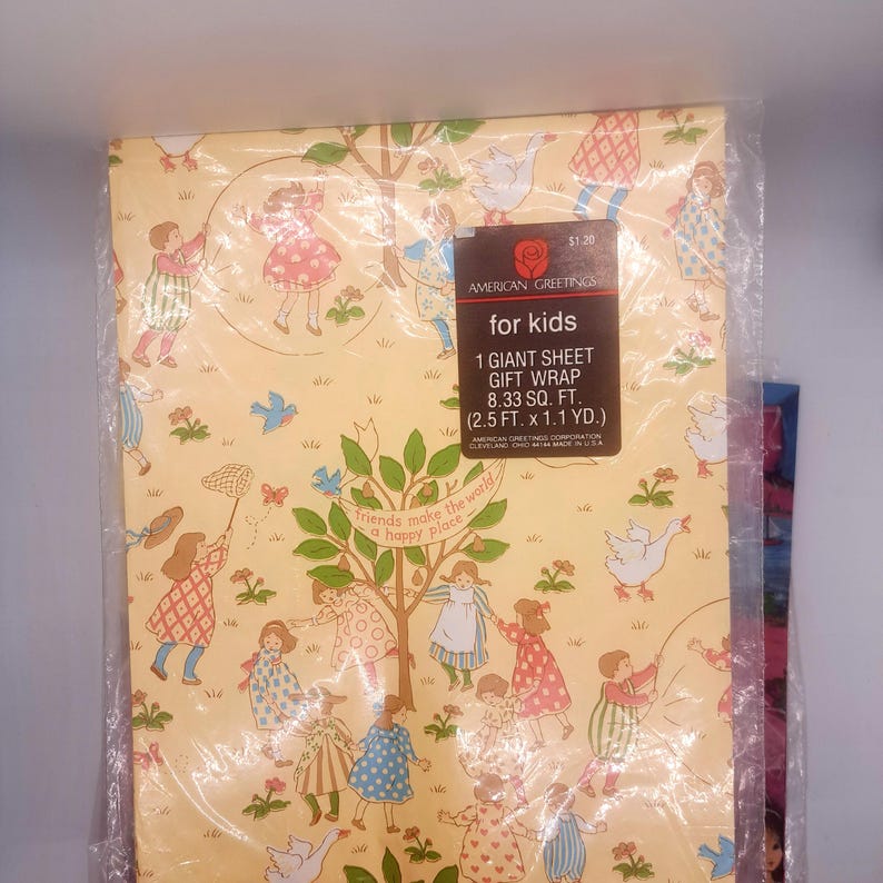 May include: A yellow sheet of gift wrap featuring a repeating pattern of children playing, trees, and geese. A black label reads: American Greetings for kids, 1 giant sheet gift wrap, 8.33 sq. ft. (2.5 ft. x 1.1 yd.), friends make the world a happy place.