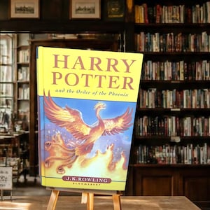 May include: A hardcover book titled Harry Potter and the Order of the Phoenix by J.K. Rowling. The cover features a yellow top section with red text and a blue bottom section displaying a large, fiery phoenix rising from flames in shades of orange.