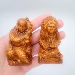May include: Two small, detailed figurines of Native American figures. One depicts a woman holding a child, the other a man in a headdress. Both are carved from a light brown material, showcasing intricate details and textures.