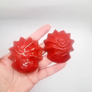 May include: Two red, star-shaped objects with a textured surface and a glossy finish. The objects have a unique, spiky design and appear to be made of a solid material. The objects are held in a hand.