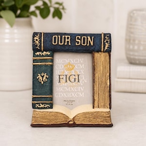 May include: A rectangular photo frame designed to look like a stack of books with a dark blue top spine labeled "OUR SON" in gold letters. The frame features a vertical green book spine on the left and an open book base, holding a 2 inch by 3 inch photo.