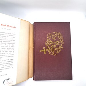 Puede incluir: Un libro de tapa dura, "The Black Mountain" de Rex Stout. La cubierta es de color burdeos oscuro con una ilustraci&oacute;n dorada. El libro est&aacute; abierto, mostrando texto en la p&aacute;gina izquierda. Publicado en enero de 1955.