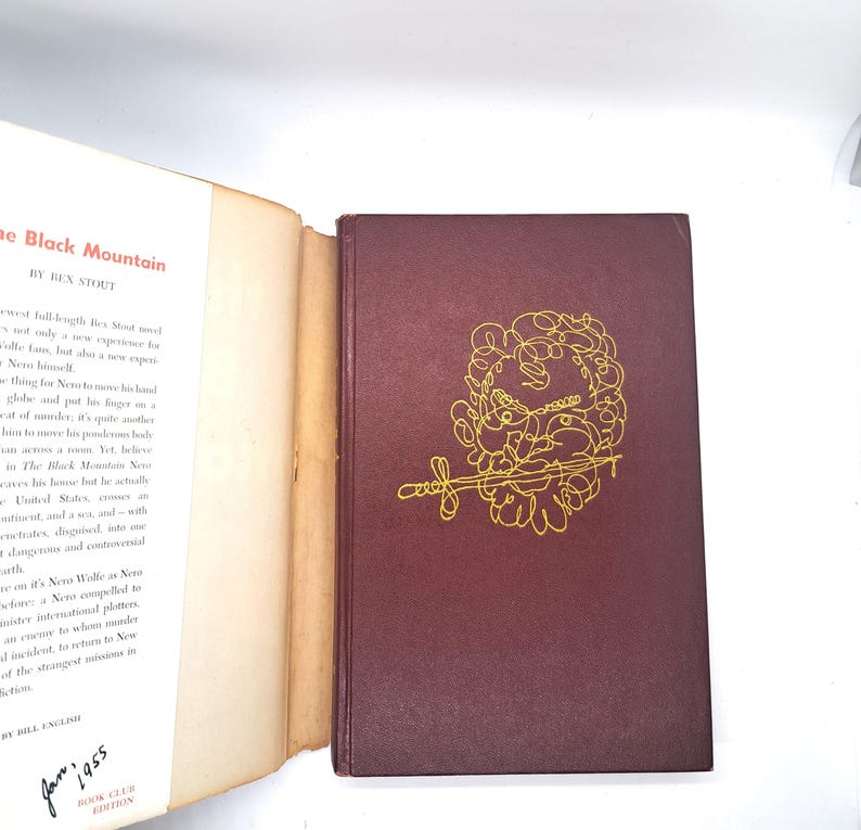 Puede incluir: Un libro de tapa dura, "The Black Mountain" de Rex Stout. La cubierta es de color burdeos oscuro con una ilustraci&oacute;n dorada. El libro est&aacute; abierto, mostrando texto en la p&aacute;gina izquierda. Publicado en enero de 1955.