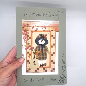 May include: A handmade craft pattern titled "Fall Memories Snowlady" featuring a snowman design. The snowman wears a plaid coat and a hat with a leaf. The pattern is on a green card with the text "Country Quilt Stitches".