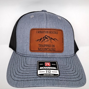 May include: A grey trucker cap with a black mesh back and a brown leather patch. The patch features a mountain graphic and the text "I RESCUE ROCKS TRAPPED IN MOUNTAINS". The cap has a curved peak and an adjustable strap.