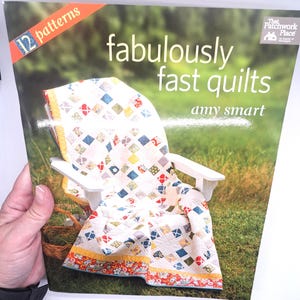 May include: A book cover titled "fabulously fast quilts" by Amy Smart. The cover features a white Adirondack chair draped with a colorful quilt. The quilt has a patchwork design with blue, red, and yellow squares. The book has "12 patterns" on the top left.